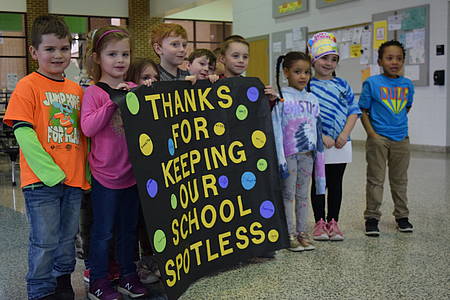 Kindergartners standing in the cafeteria present a class-made poster to the custodial staff. It's decorated with large, colorful spots and read, "Thanks for keeping our school spotless."